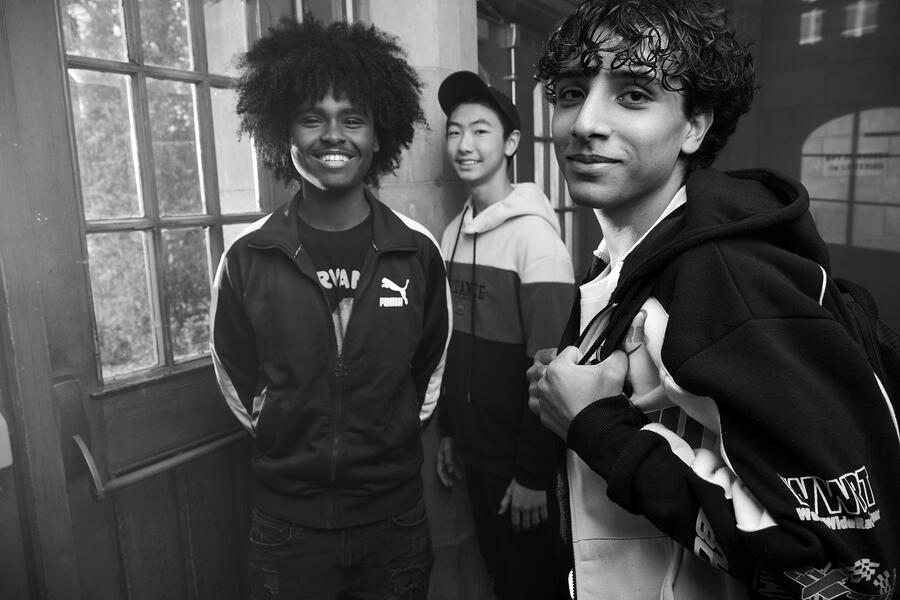 three boys standing in the front hall of their school with backpacks, smiling at the camera. 